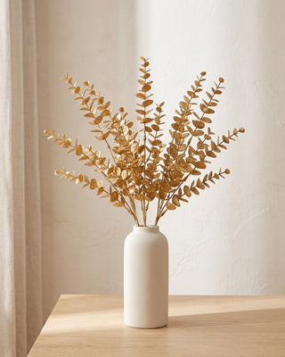 Artificial gold leaf stems standing in a plain off-white vase on a pale oak table; no vase is included