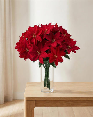 A red artificial poinsettia bouquet in a clear glass vase on a side table; vase not included