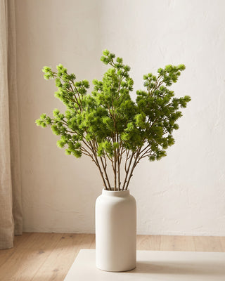 An artificial pine branch standing in a plain off-white vase on a pale oak table; each order is one branch, and no vase is included