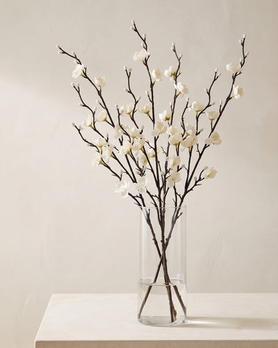 Five artificial white plum blossom stems in a plain clear glass vase on a pale surface. Vase not included.