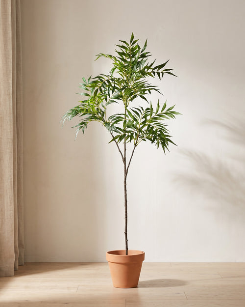 Artificial bamboo branch with narrow lance-shaped leaves, standing in a plain terracotta pot on a pale oak floor (pot not included).