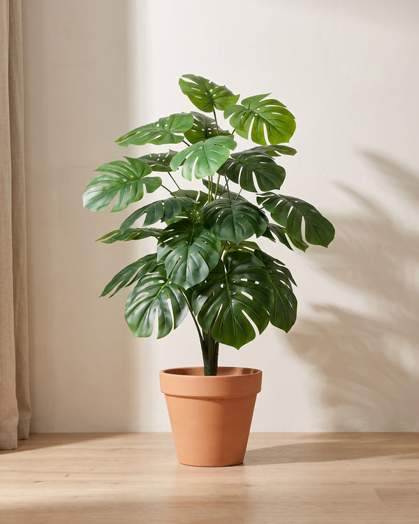 Artificial monstera bush in a plain terracotta pot against a warm off-white plaster wall