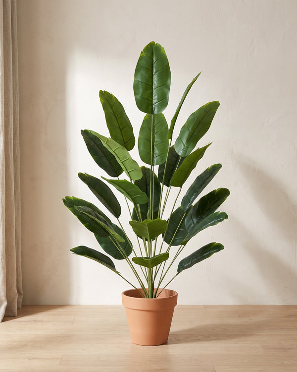 Artificial banana leaf bush in a plain terracotta pot against a warm off-white plaster wall