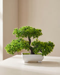 Artificial flowering bonsai in its plain white molded pot on a pale surface, canopy wider than the tree is tall