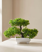 Artificial flowering bonsai in its plain white molded pot on a pale surface, canopy wider than the tree is tall