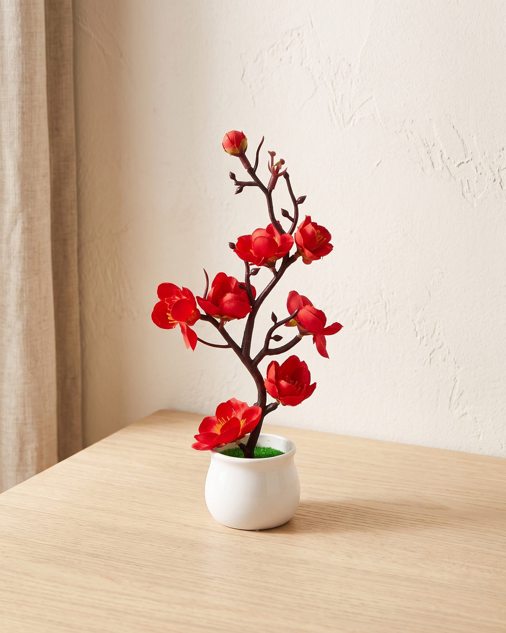 Artificial red plum blossom bonsai in a white ceramic pot on a pale oak table