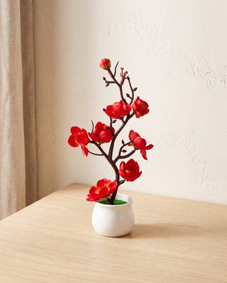 Artificial red plum blossom bonsai in a white ceramic pot on a pale oak table