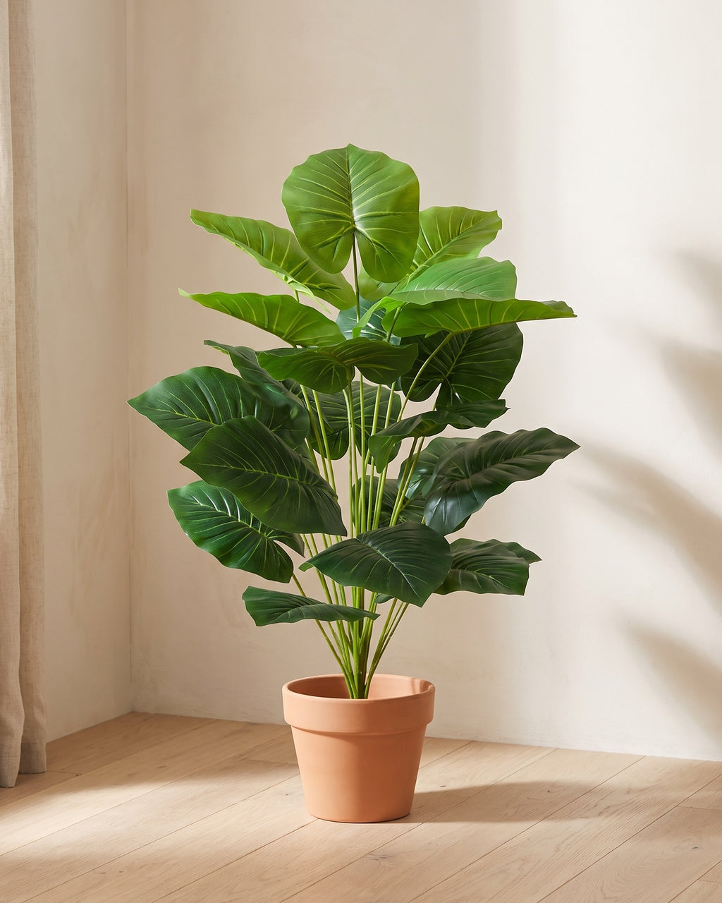 The 39.4 in artificial elephant ear plant in the corner of a bare room, shown in a plain terracotta pot (pot not included).