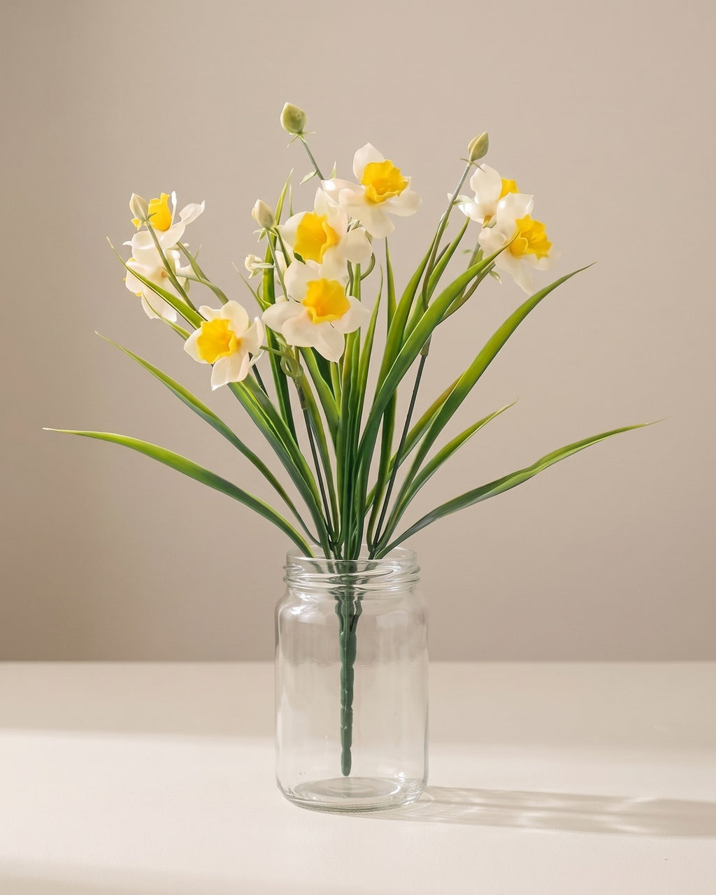 One bunch of artificial daffodils in a plain clear glass jar on a pale surface. Jar not included.