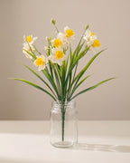 One bunch of artificial daffodils in a plain clear glass jar on a pale surface. Jar not included.