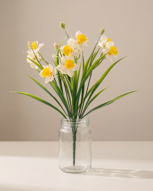 One bunch of artificial daffodils in a plain clear glass jar on a pale surface. Jar not included.