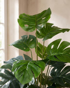 Close view of the artificial monstera's split, perforated leaves and slim stems in soft window light.