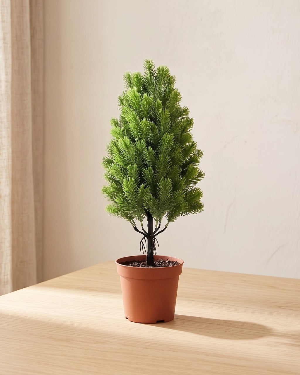 A tall narrow artificial mini Christmas tree with soft pine needles, in a plain nursery pot on a pale oak table; pot not included