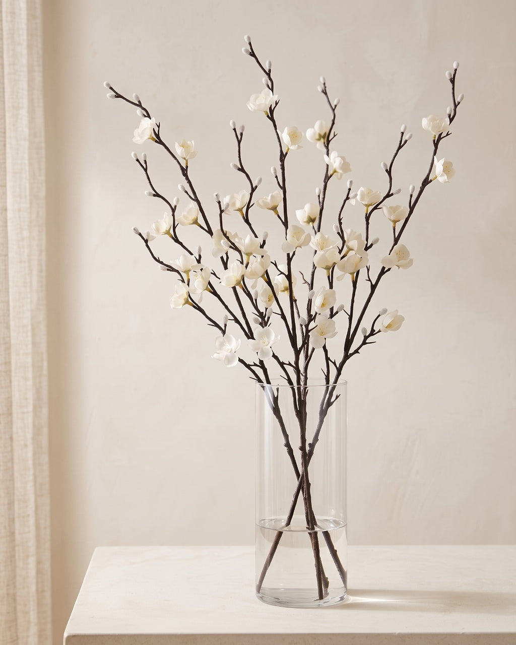 White artificial plum blossom stems standing in a vase on a pale oak table; no vase is included
