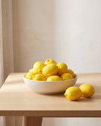 A plain shallow bowl of artificial lemons on a pale oak table, two beside it, in a bright room