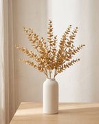 Artificial gold leaf stems standing in a plain off-white vase on a pale oak table; no vase is included