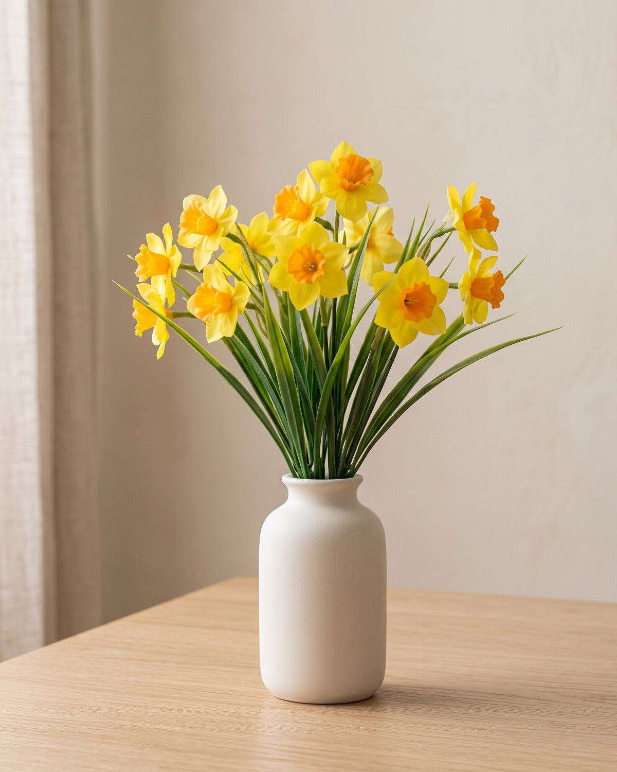 Orange and yellow artificial daffodil bunch standing in a plain off-white vase on a pale oak table; no vase is included
