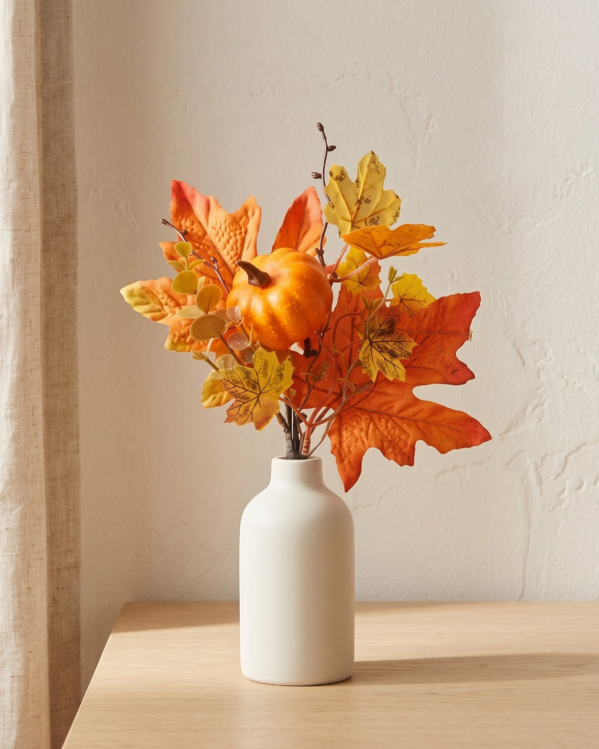 Artificial fall pick of maple leaves, small pumpkins and yellow leaves, in a plain off-white bud vase on a pale oak tabletop, vase not included