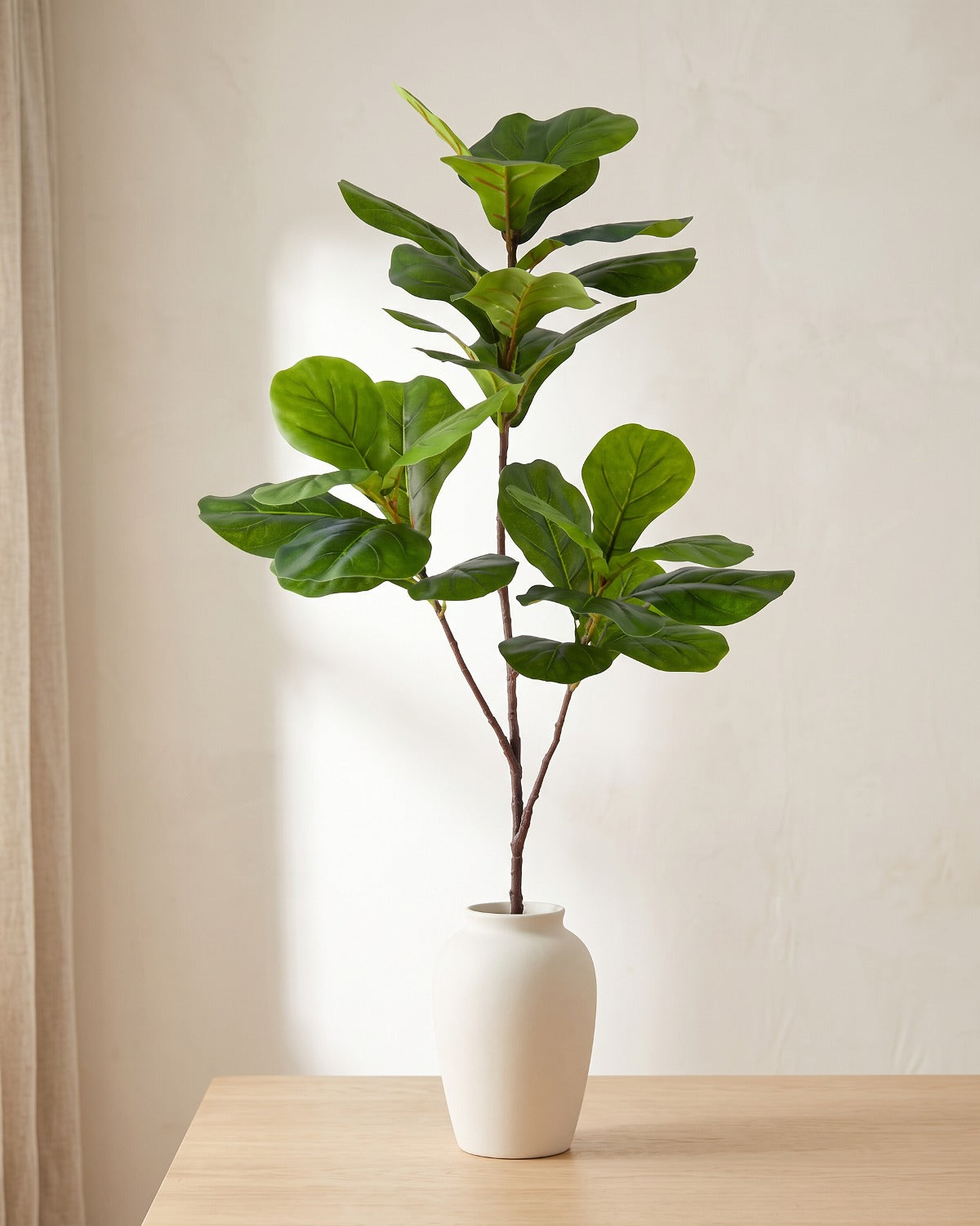 An artificial tree branch standing in a plain off-white vase on a pale oak table; each order is cut stems, and no vase is included