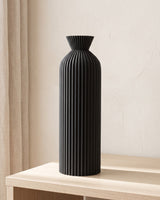 A tall black ribbed bottle vase, empty, on a pale oak console