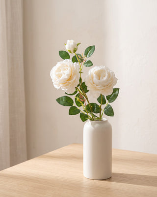 An artificial rose branch standing in a plain off-white vase on a pale oak table; each order is one branch, and no vase is included