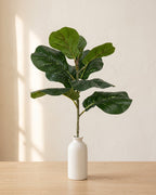A short artificial fiddle leaf fig branch, a single stem of broad glossy leaves, in a plain off-white bud vase on a pale oak table; vase not included
