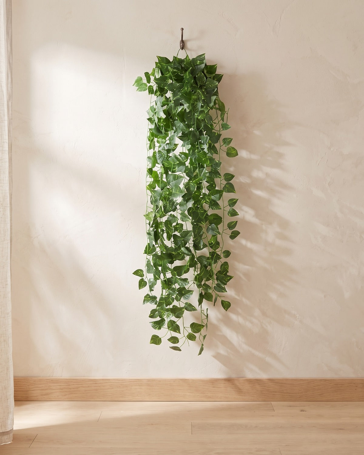Pointed leaf artificial ivy garland hanging from a simple hook on an off-white plaster wall, its full drop clear of the floor