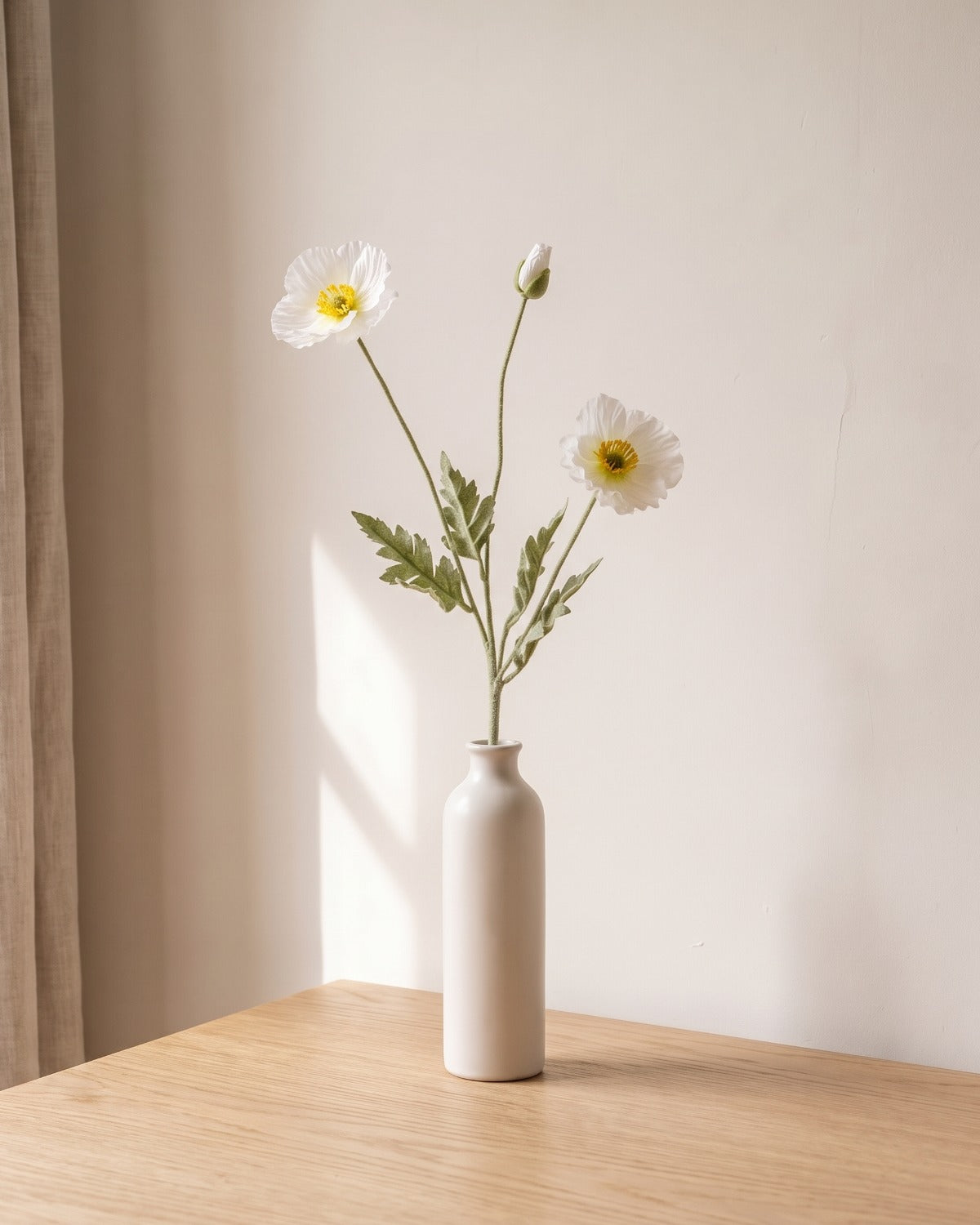 A white artificial poppy stem, one stem with two open flowers and a bud, in a plain off-white bud vase on a pale oak table; vase not included