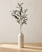 Artificial olive branch with dark olives along the stem, in a plain off-white bud vase on a pale oak tabletop, vase not included