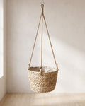 A woven hanging plant basket on jute ropes, empty, against a plaster wall