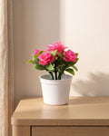A small artificial flowering pot with bright pink blooms, in the white pot it comes in, on a pale oak table