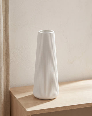 A tall fluted tapered vase in matte white, empty, on a pale oak console