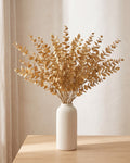 Twenty artificial gold leaf stems standing in a plain off-white vase on a pale oak table; no vase is included