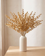 Twenty artificial gold leaf stems standing in a plain off-white vase on a pale oak table; no vase is included