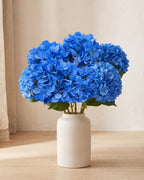 Blue artificial hydrangea flowers standing in a plain off-white vase on a pale oak table; no vase is included