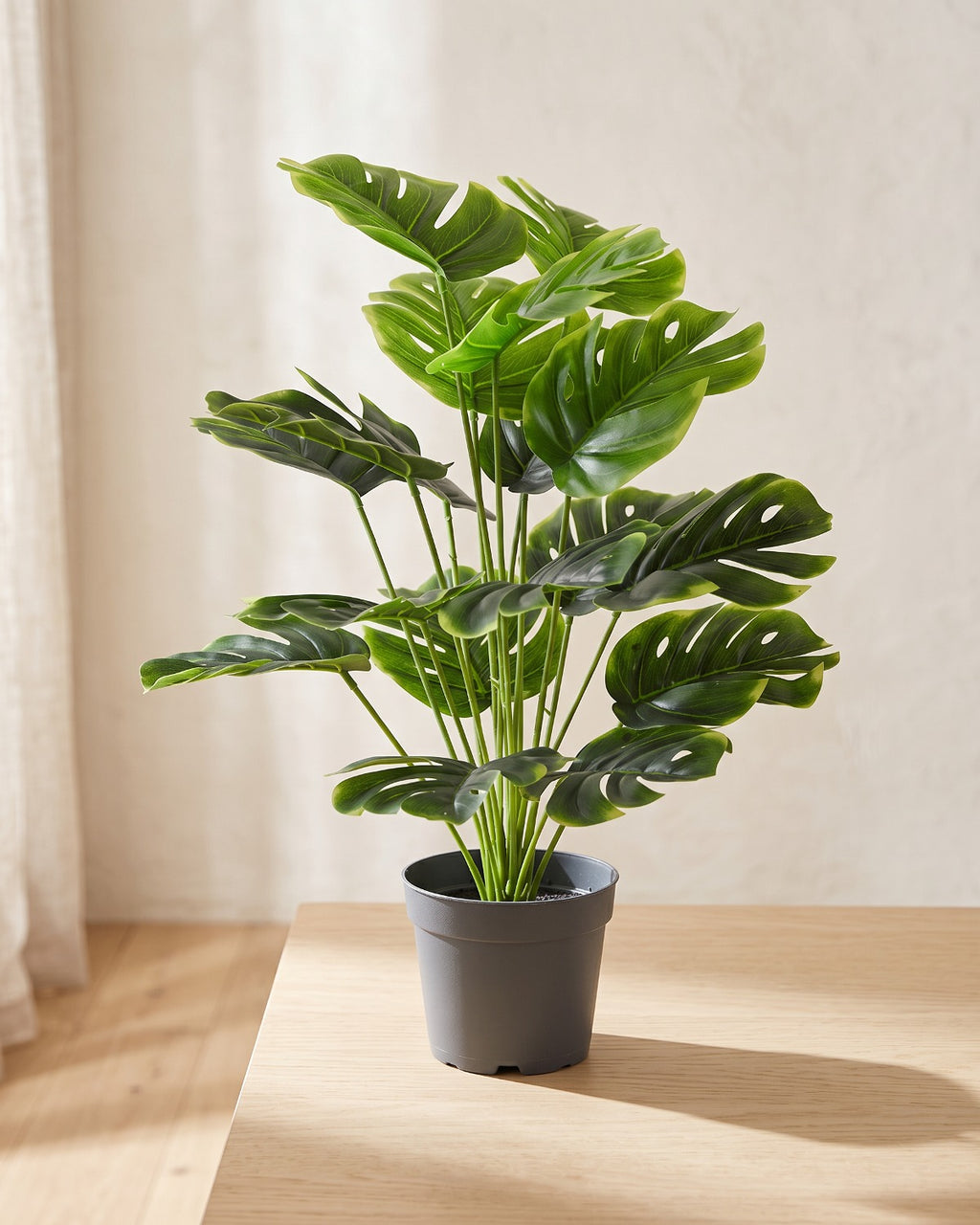 An artificial monstera plant in a plain nursery pot on a pale oak table in a bright room; pot not included