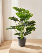 An artificial monstera plant in a plain nursery pot on a pale oak table in a bright room; pot not included