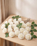 Twenty-five ivory foam rose heads on wire stems with green leaf sprays, laid out on a pale oak table