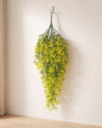 Yellow artificial wisteria vine hanging from a simple hook on an off-white plaster wall, its full drop clear of the floor