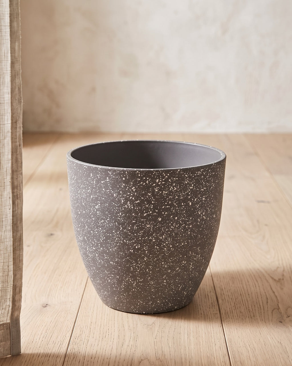 Stone stone effect plant pot standing empty on pale oak floorboards against an off-white plaster wall