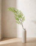 A single artificial asparagus fern frond, fine and feathery, in a plain off-white floor vase on pale oak floorboards (vase not included).