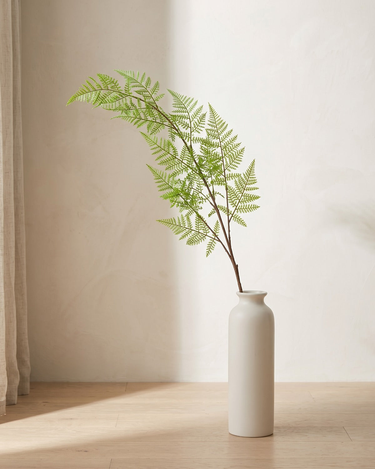 A single artificial asparagus fern frond, fine and feathery, in a plain off-white floor vase on pale oak floorboards (vase not included).