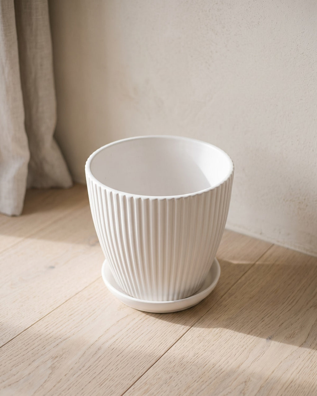 A white fluted plant pot and its saucer, empty, on pale oak floorboards