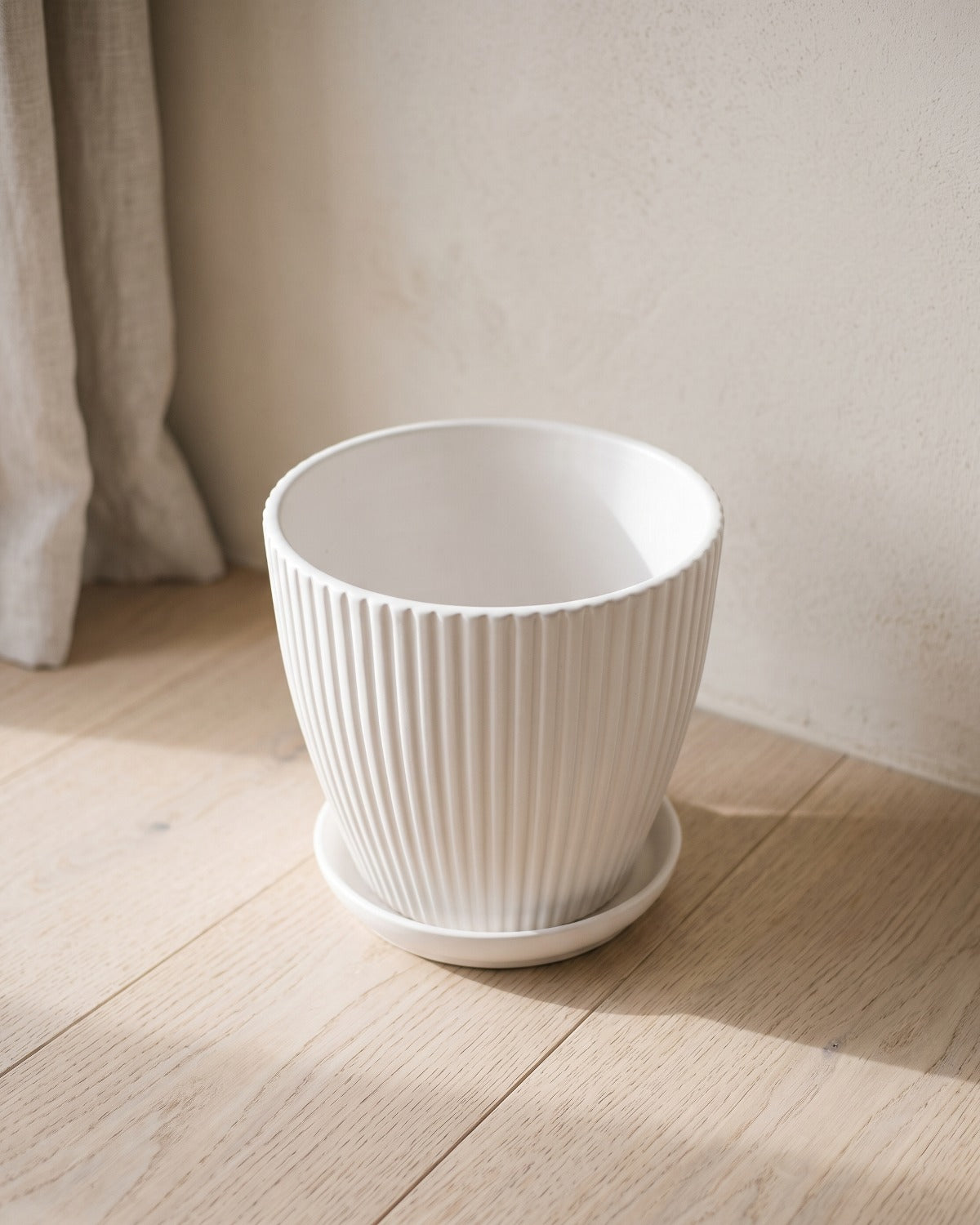 A white fluted plant pot and its saucer, empty, on pale oak floorboards