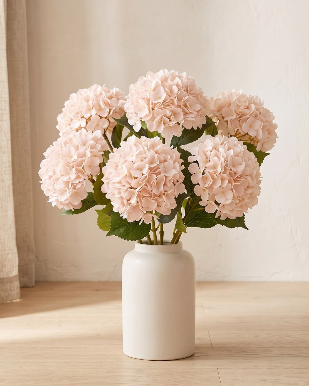 Light Pink artificial hydrangea flowers standing in a plain off-white vase on a pale oak table; no vase is included