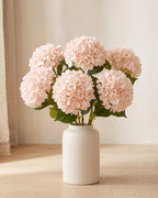 Light Pink artificial hydrangea flowers standing in a plain off-white vase on a pale oak table; no vase is included