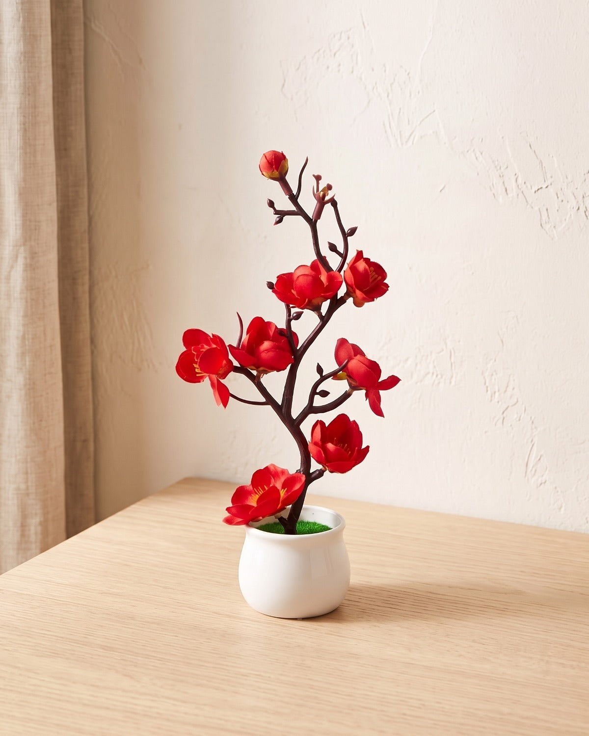 A small artificial plum blossom bonsai in scarlet red bloom, in the white ceramic pot it comes in, on a pale oak table