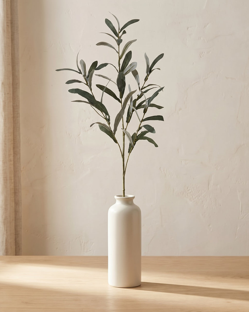 Artificial olive branch with silvery green leaves and no olives, in a plain off-white bud vase on a pale oak tabletop, vase not included
