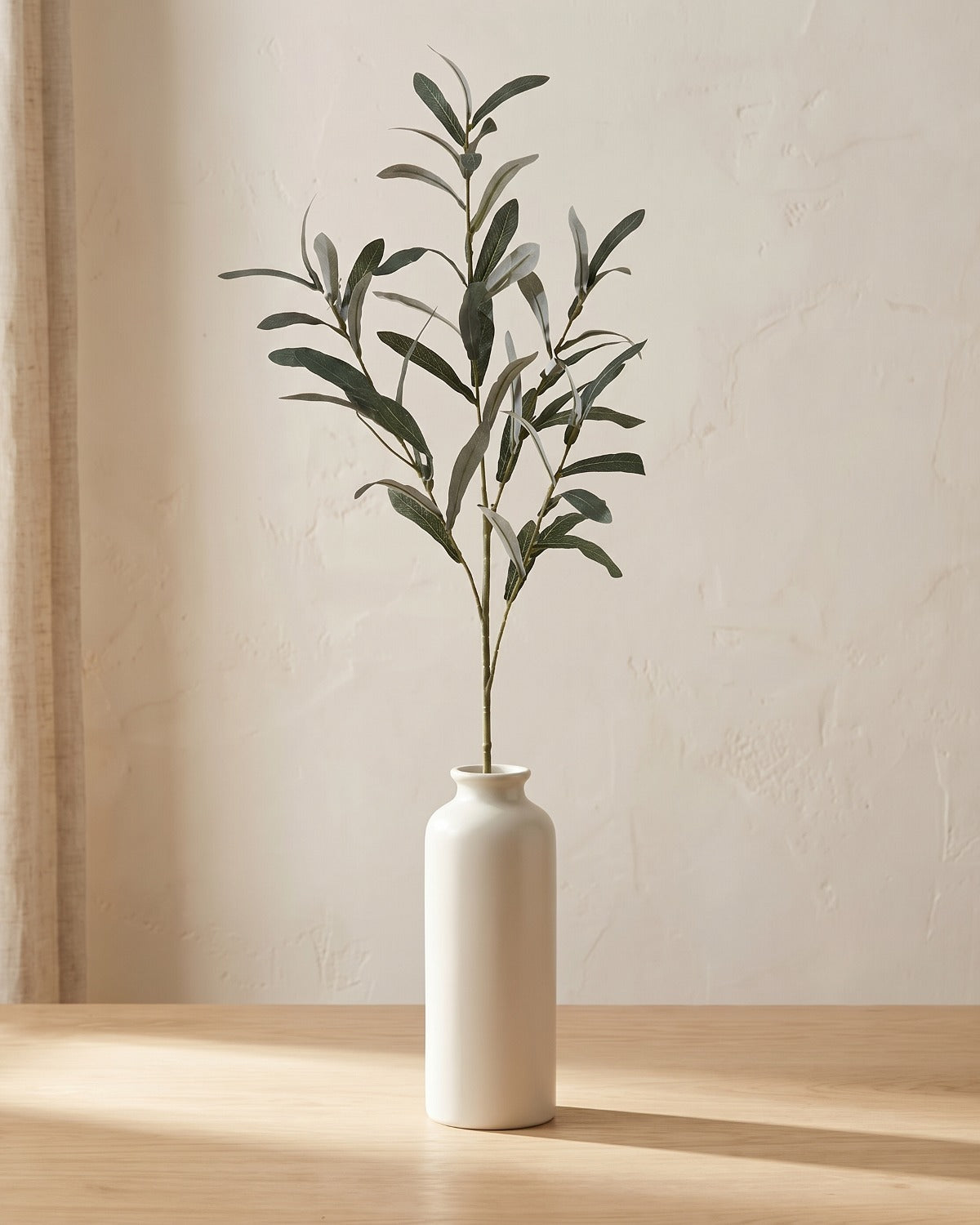 Artificial olive branch with silvery green leaves and no olives, in a plain off-white bud vase on a pale oak tabletop, vase not included