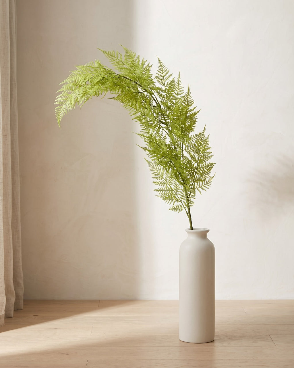 A single artificial plumosa fern frond, dense and plume-like, in a plain off-white floor vase on pale oak floorboards (vase not included).